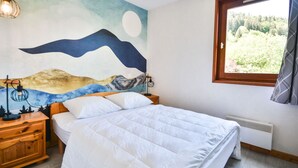 A bedroom with a bed, a nightstand, a wall-mounted lamp, a window with a view of greenery, and a wall mural. - Central Morzine apartment, 6 pers, south balcony (Morzine)