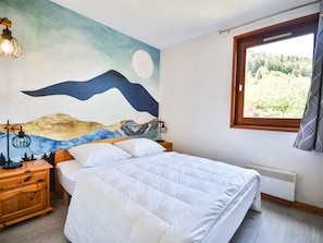 A bedroom with a bed, a nightstand, a wall-mounted lamp, a window with a view of greenery, and a wall mural. - Central Morzine apartment, 6 pers, south balcony (Morzine)