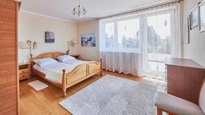 A bedroom with a wooden bed, a desk, a chair, and large windows with sheer curtains. - Holiday Home in Swinoujscie Near Baltic Beach (Swinoujscie)