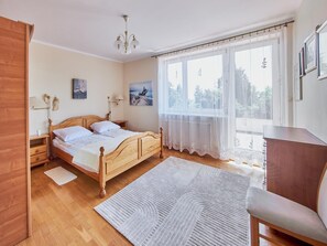 Une chambre à coucher avec un lit en bois, une table de chevet, une commode, une chaise, une grande fenêtre avec des rideaux légers et un luminaire au plafond.