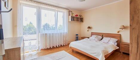 A bedroom with a bed, bedside tables, a nightstand, a window with curtains, and a view of greenery outside.