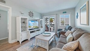 A living room with a television, white entertainment unit, grey sofa, and a view of the outdoors through a glass door. - Gulf Shores Plantation 2123 Fun & Games (Gulf Shores)
