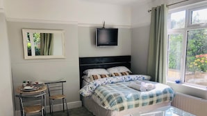 A neatly arranged bedroom with a bed, a small table with chairs, a TV mounted above the bed, and a window with curtains. - The studio (St Austell)