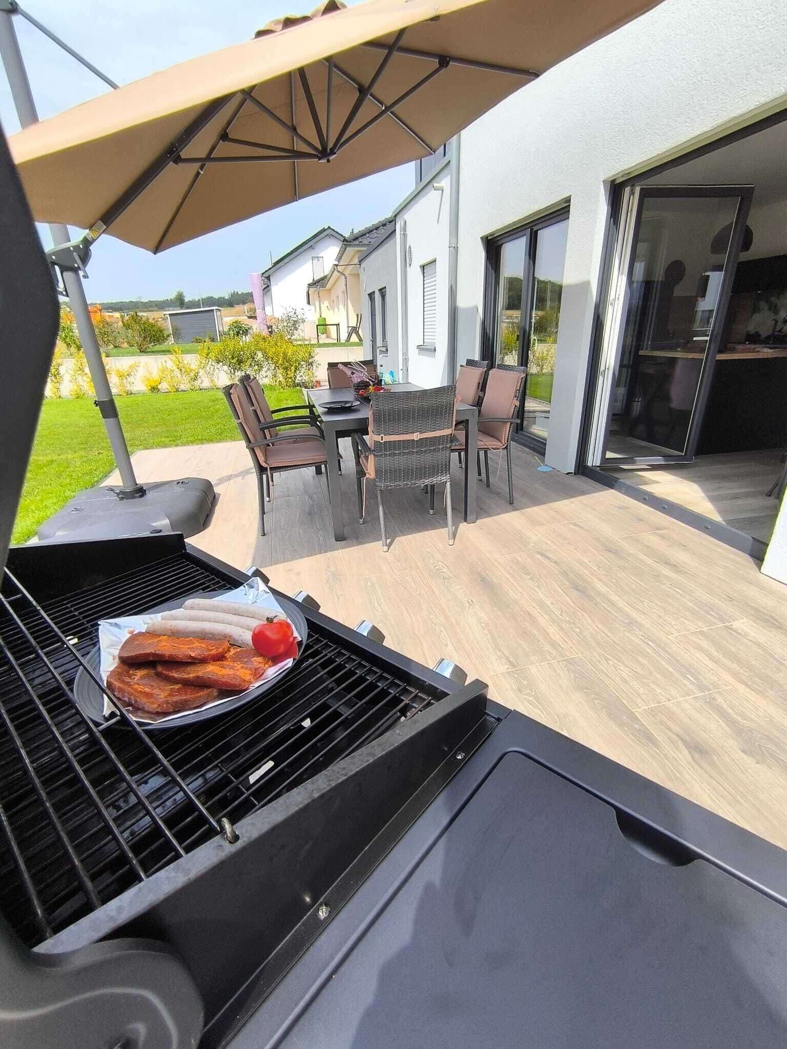A barbecue grill with sausages and a tomato on a patio with outdoor furniture and a house in the background.