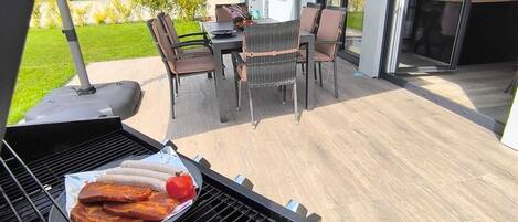 A barbecue grill with sausages and a tomato on a patio with outdoor furniture and a house in the background.