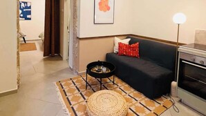 Una sala de estar con un sofá oscuro, un otoman tejido redondo, una mesita auxiliar negra pequeña, una lámpara de pie moderna y una cocina con estufa.