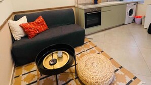 Un espacio habitable compacto con sofá, una mesita de centro, un otoman tejido, una zona de cocina con lavadora y una ventana.