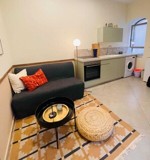 A compact living space with a sofa, a small coffee table, a woven ottoman, a kitchen area with a washing machine, and a window. - Charming 1BR in Nachlaot  Steps to the Shuk (Jerusalem)