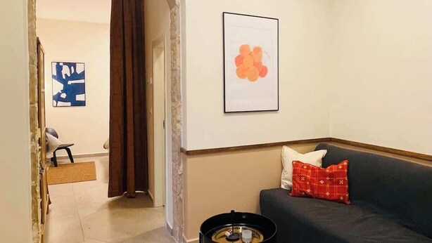 Una sala de estar con un sofá oscuro, un otoman tejido redondo, una mesita auxiliar negra pequeña y un tapete estampado. Hay un cuadro en la pared y un vano abierto que da a otra habitación.