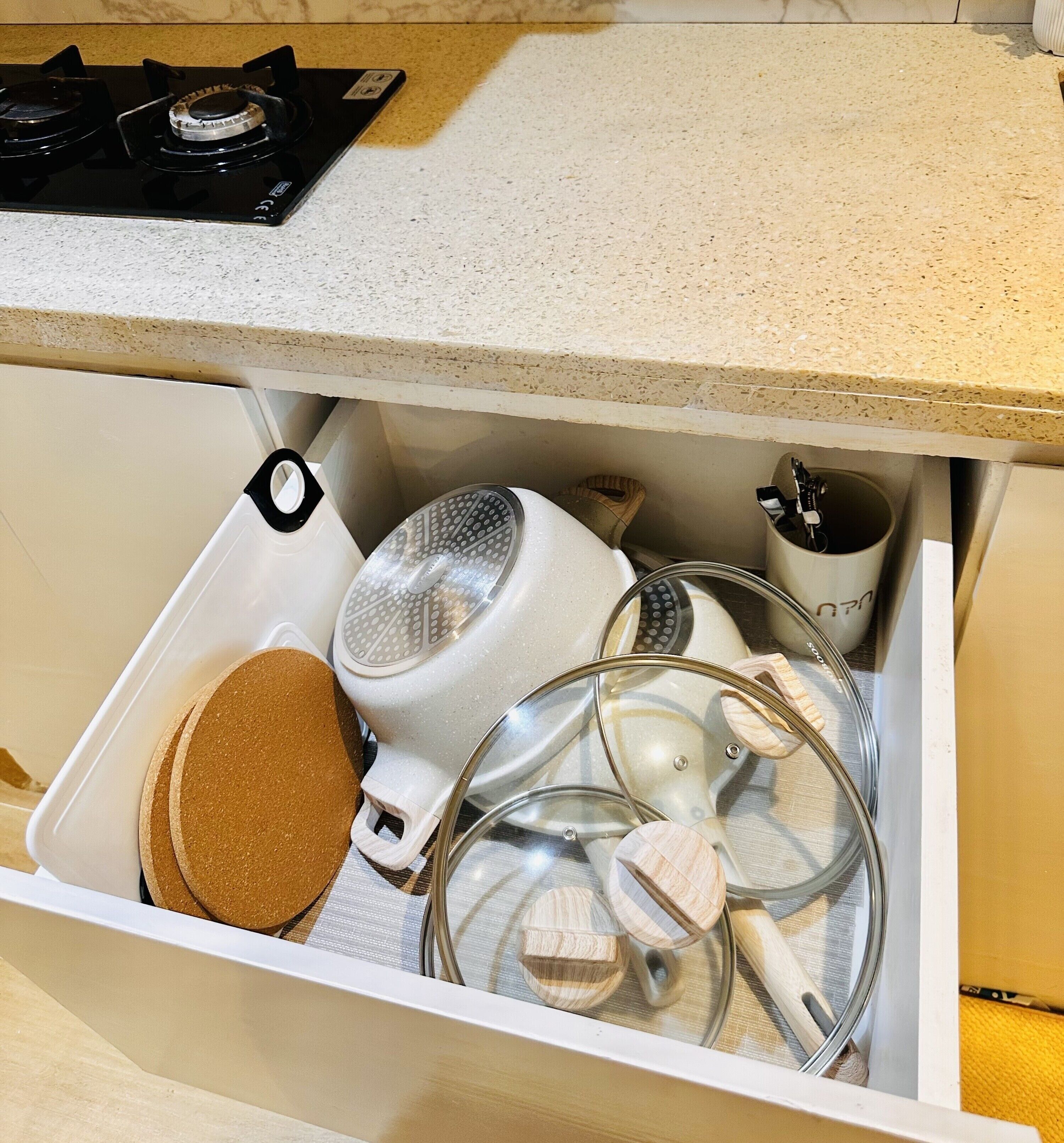 A kitchen drawer with a white countertop above, containing a white kettle, a glass container, and a wooden spoon.