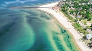 An aerial view of a coastal area with clear turquoise waters, a sandy beach, and a residential area with houses and roads. - Pier Cove Estate - Pool, Pickleball, Hot Tub, Firepit - Sleeps up to 23 (Fennville)