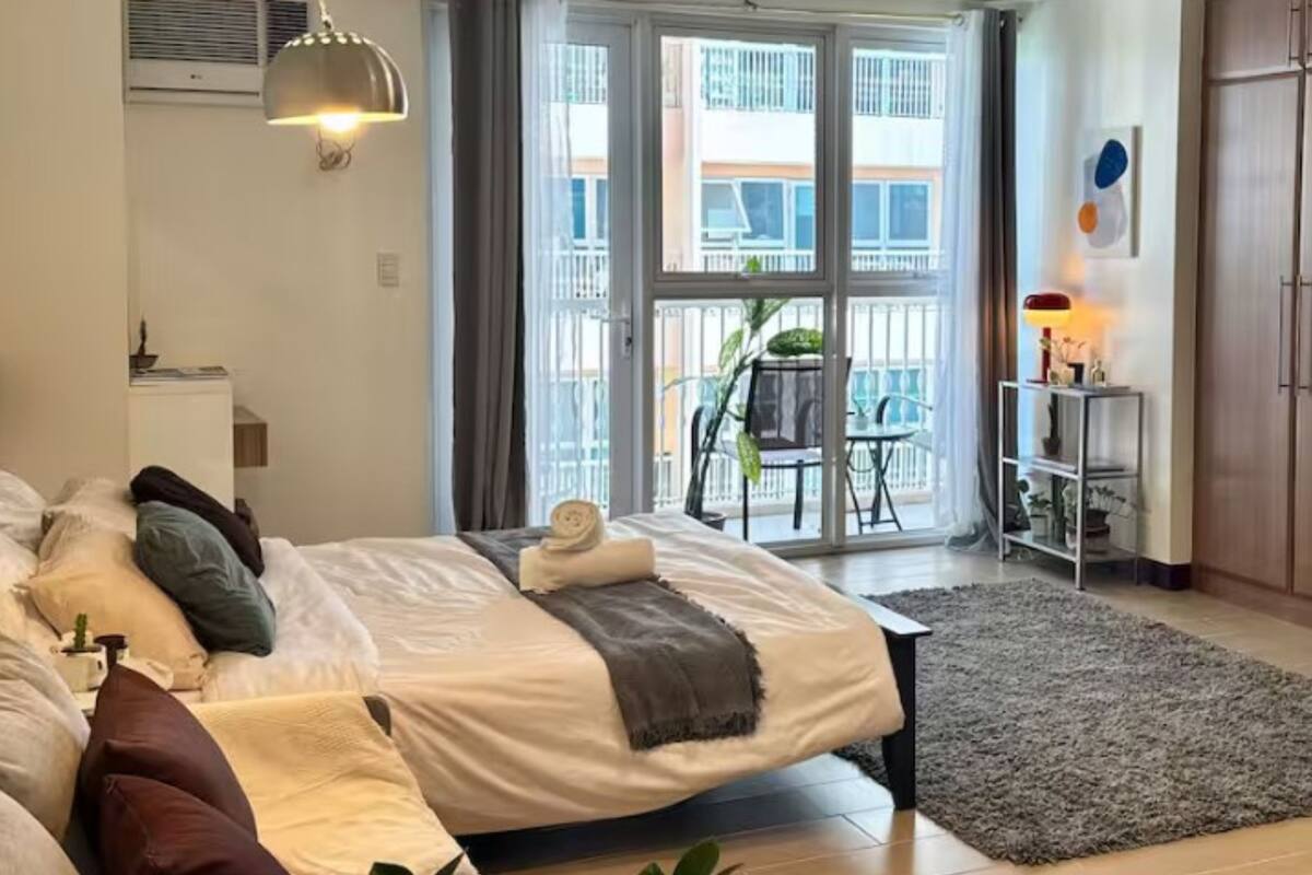 A bedroom with a bed, a desk, a chair, a balcony, and a view of buildings.