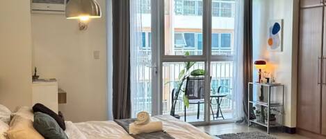 A bedroom with a bed, a desk, a chair, a balcony, and a view of buildings.