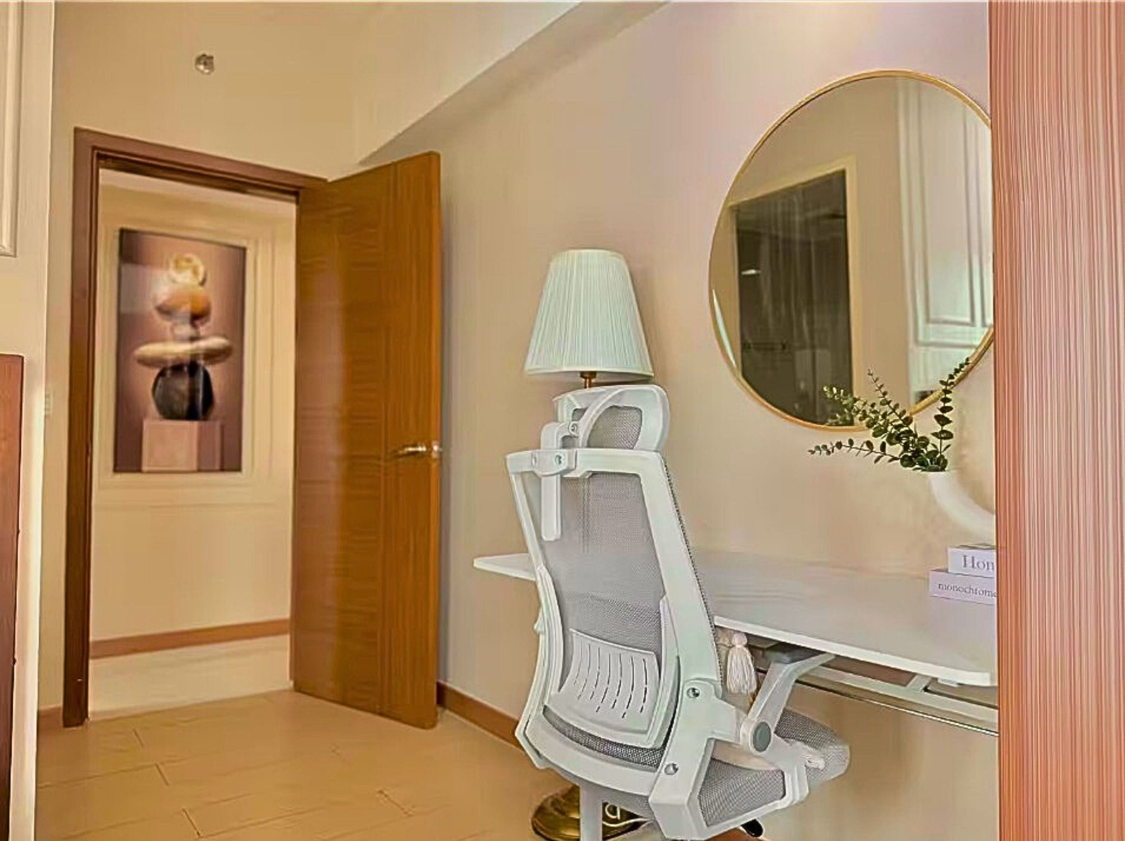 A room with a wooden door, a white desk with a lamp, a chair, and a round mirror.