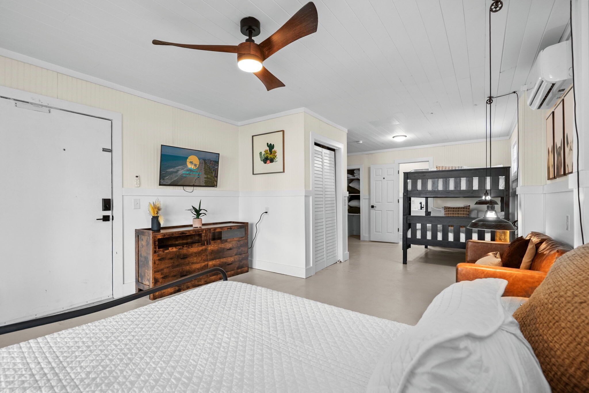 A bedroom with a bed, a wooden dresser, a ceiling fan, and a TV on the wall.