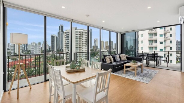 A spacious living room with a large window, a black sofa, a dining table, and a view of tall buildings. - Skyline Serenity - Modern Escape + Stunning Views (Surfers Paradise)