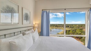 A bedroom with a large bed, a ceiling fan, and a window with a view of a lake and trees.