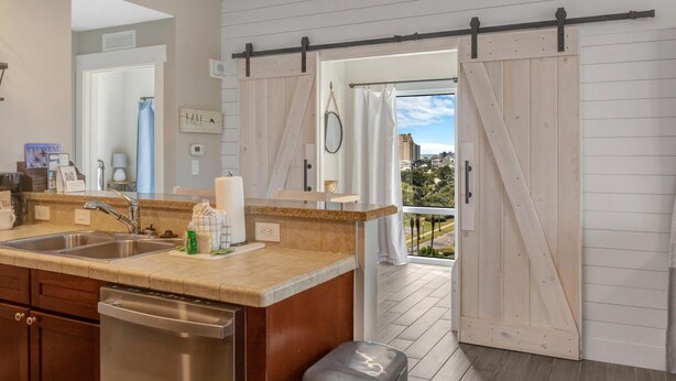 A kitchen with a stainless steel dishwasher, a sink, and a countertop with a towel and a bottle. There is a sliding door leading to a balcony with a view of buildings and trees.