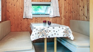 Une pièce chaleureuse aménagée comme un chalet, avec un intérieur en bois, une petite table recouverte d’une nappe à motifs, deux bancs rembourrés, une fenêtre avec des rideaux et un luminaire rouge suspendu.