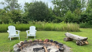 A fire pit surrounded by rocks and logs, with two white Adirondack chairs in the background.