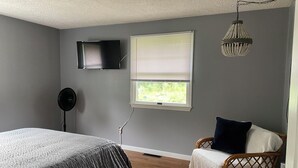 A bedroom with a bed, a chair, a TV, a window with blinds, and a hanging light fixture.