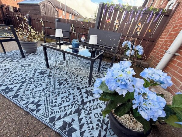 An outdoor patio with a patterned rug, a table with chairs, and a potted plant with blue flowers. - 3BR Modern House, Garden, Parking, Sleeps 6, Wifi (Strabane)
