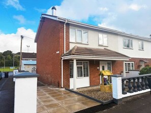 A two-story house with a brick facade, a white front door, and a small porch with a garden bed. - 3BR Modern House, Garden, Parking, Sleeps 6, Wifi (Strabane)