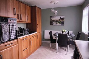 A kitchen with wooden cabinets, a dining table with chairs, a hanging light fixture, and a cityscape painting on the wall. - 3BR Modern House, Garden, Parking, Sleeps 6, Wifi (Strabane)