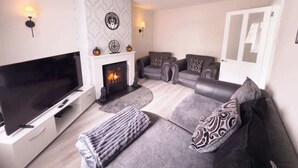 A living room with a flat-screen TV, a fireplace, grey sofas, and patterned cushions. - 3BR Modern House, Garden, Parking, Sleeps 6, Wifi (Strabane)