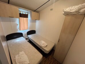 A compact room with two beds, a small window with curtains, a wardrobe, and a bedside table with a lamp. - Air-conditioned mobile home with a terrace—between the sea and the Camargue (Lunel)