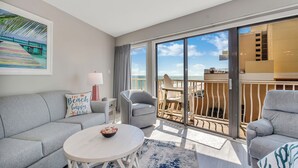 A modern hotel room with a balcony, a sofa, a round coffee table, and a painting of a beach scene.