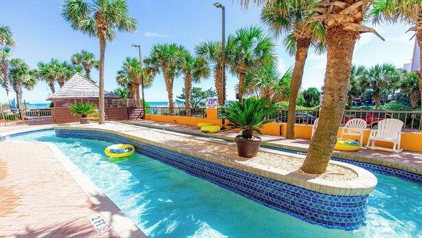 A swimming pool with a tiled edge, palm trees, and a beach in the background.