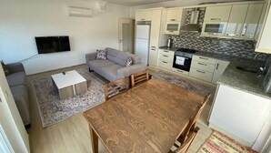 A compact living space with a sofa, a dining table, a kitchen area with a stove, and a TV mounted on the wall. - Mitra apart (Antalya)