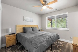 Un dormitorio con cama, mesita de noche, ventilador de techo, ventana con persianas y una obra de arte colgada en la pared.