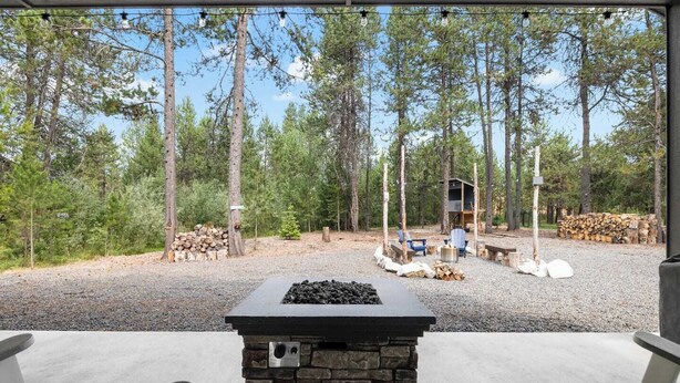 A covered outdoor area with a stone fire pit, two Adirondack chairs, and a small wooden structure in a forested setting.