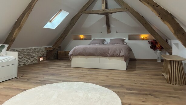 Et soveværelse med skråt loft, en seng med gavl, en stenvæg, trægulv, en hvid kommode, et lille rundt bord og et ovenlys.