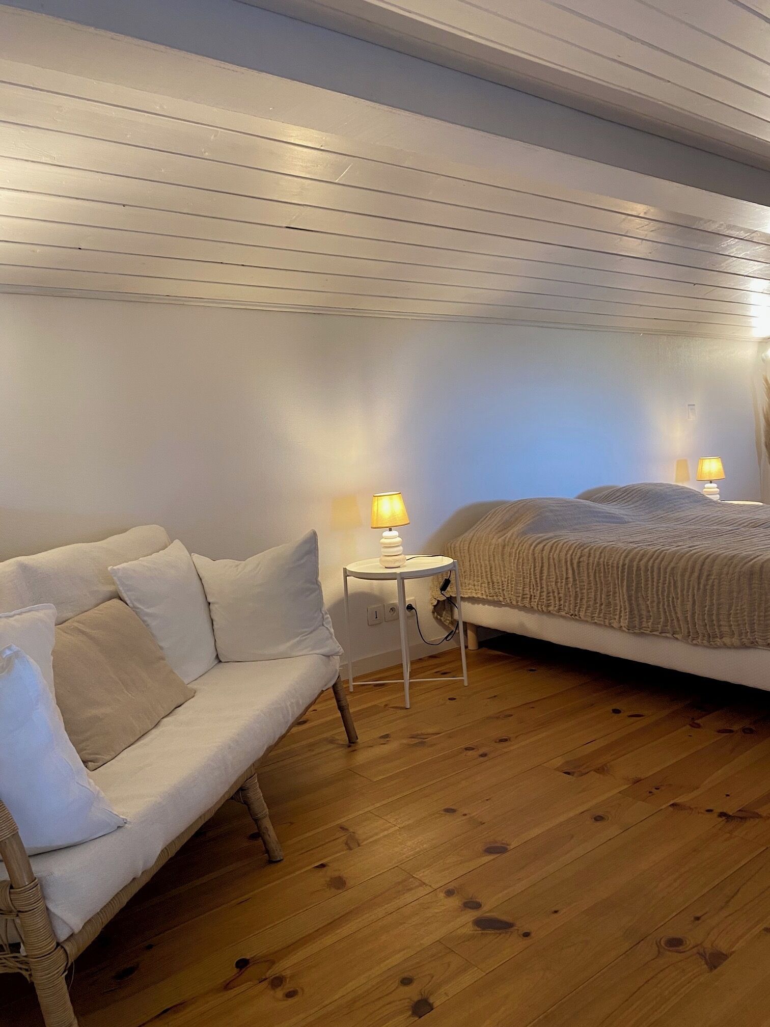A room with a white sofa, a bed with a beige blanket, and two bedside lamps on a small white table.