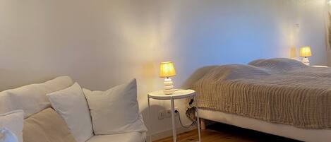 A room with a white sofa, a bed with a beige blanket, and two bedside lamps on a small white table.