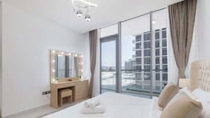 A modern bedroom with a large bed, a vanity with a mirror and lights, and a view of a building through large windows.