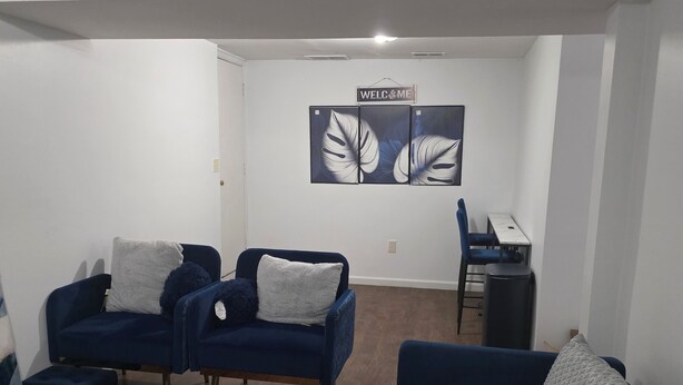 A modern living room with two blue sofas, a white coffee table, and a black rug. There are two framed artworks on the wall and a small desk with a chair.
