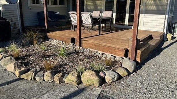 A covered patio area with a wooden deck, stone border, and outdoor furniture.