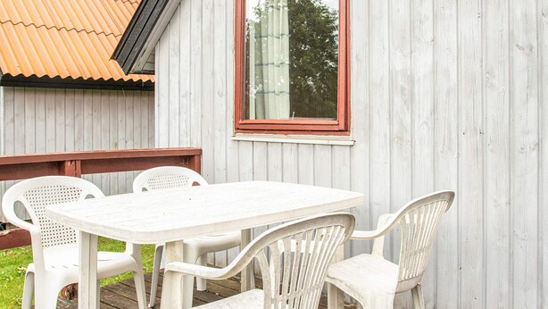 Et sæt hvide plastborde og stole på en træterrasse med et bølgebliktag og en trævæg med et vindue.