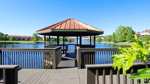 A wooden deck leading to a gazebo over a body of water, surrounded by greenery and buildings. - Bright 3B w Pool Close to Disney Encantada 8545 (Kissimmee)