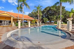 A swimming pool with a tiled deck, palm trees, and a building with a red roof in the background. - Bright 3B w Pool Close to Disney Encantada 8545 (Kissimmee)