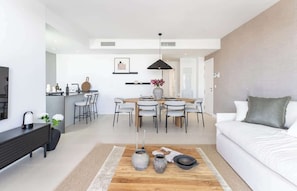 Una sala de estar moderna con un sofá, una mesa de centro con adornos, un comedor con mesa y sillas, y una cocina con mostrador y electrodomésticos.