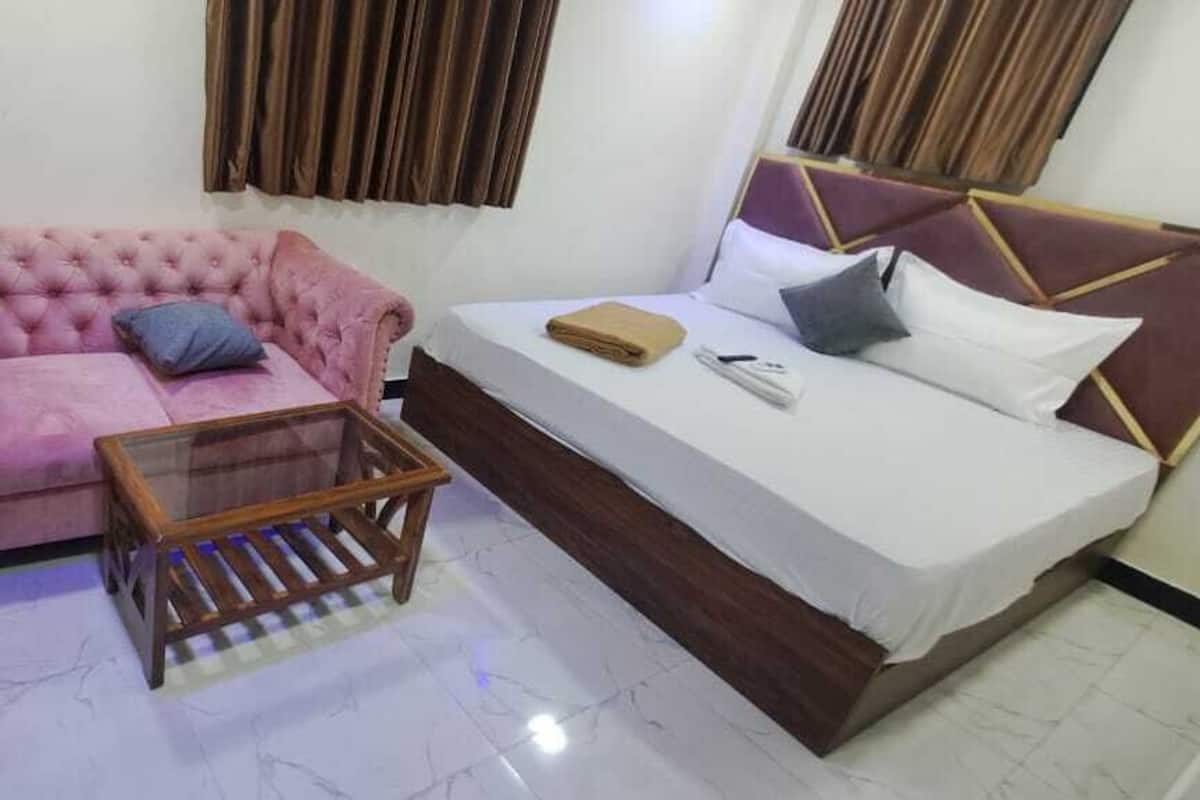 A room with a pink sofa, a wooden side table, a bed with a headboard, and a small wooden stool.