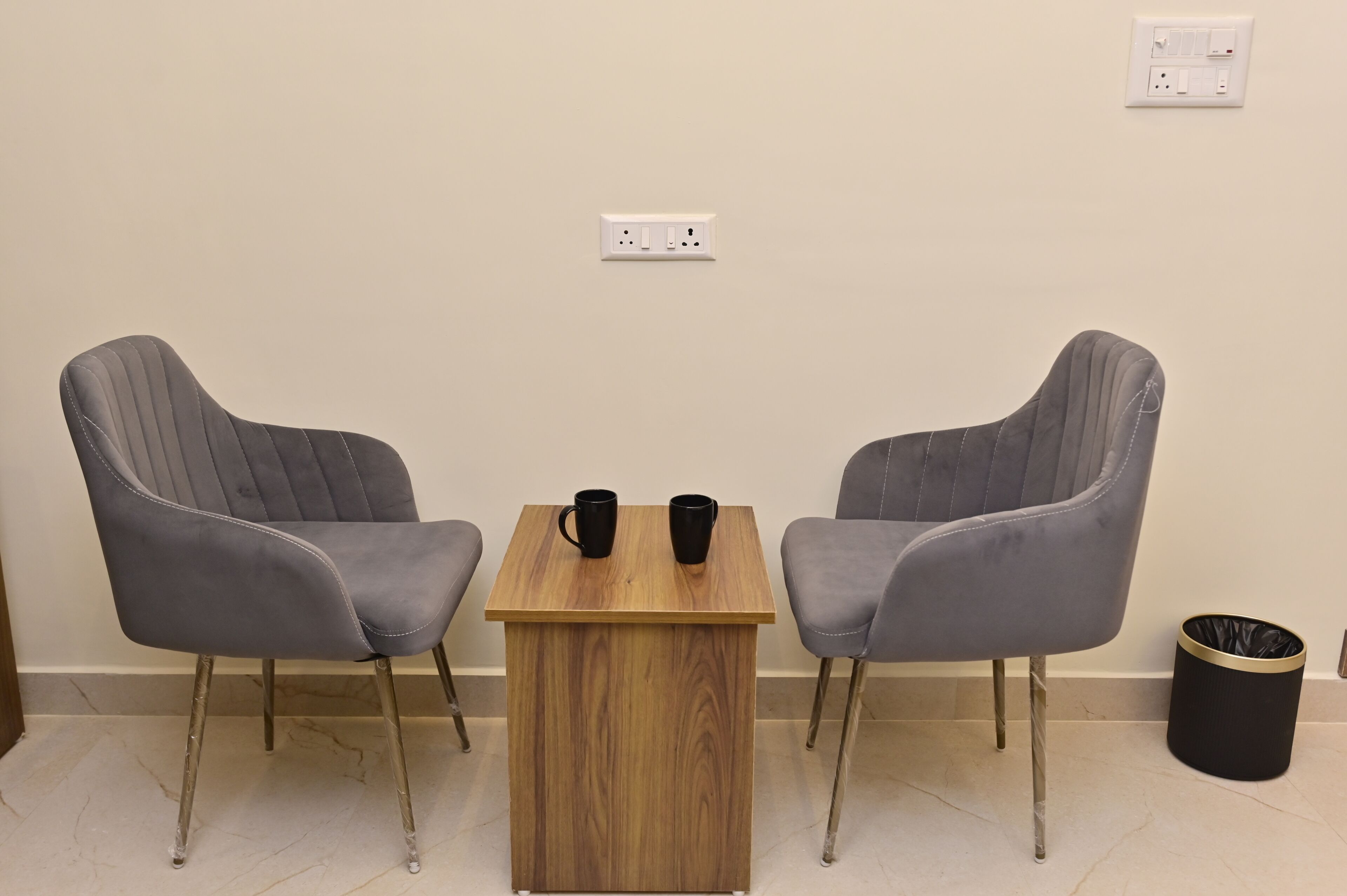 A small room with two grey chairs, a wooden table, and a black trash bin.