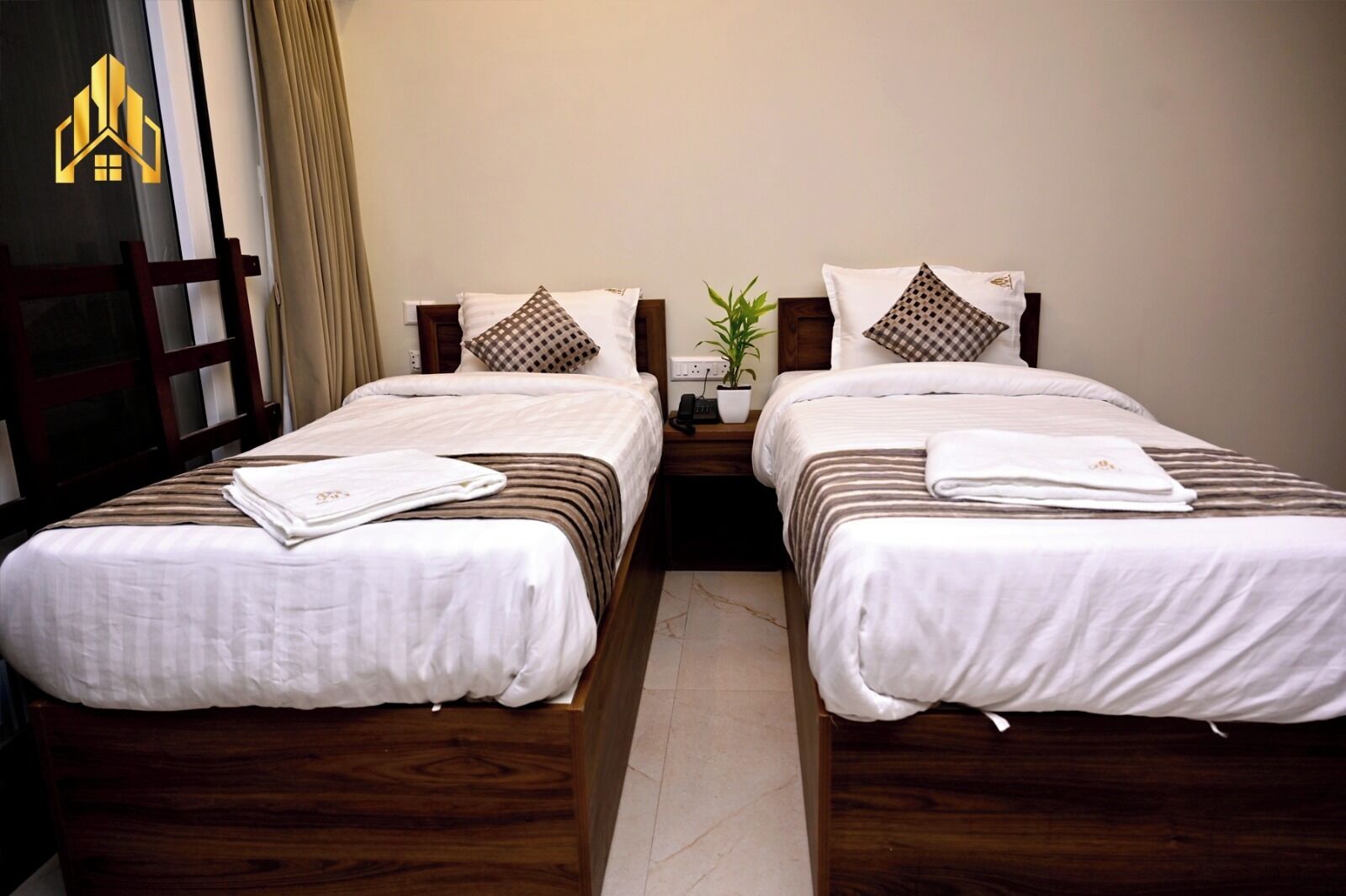 Une chambre d’hôtel avec deux lits, chacun doté d’une tête de lit en bois, de draps blancs et d’un oreiller à carreaux. Une table de chevet avec une lampe et une plante en pot se trouve entre les deux lits.