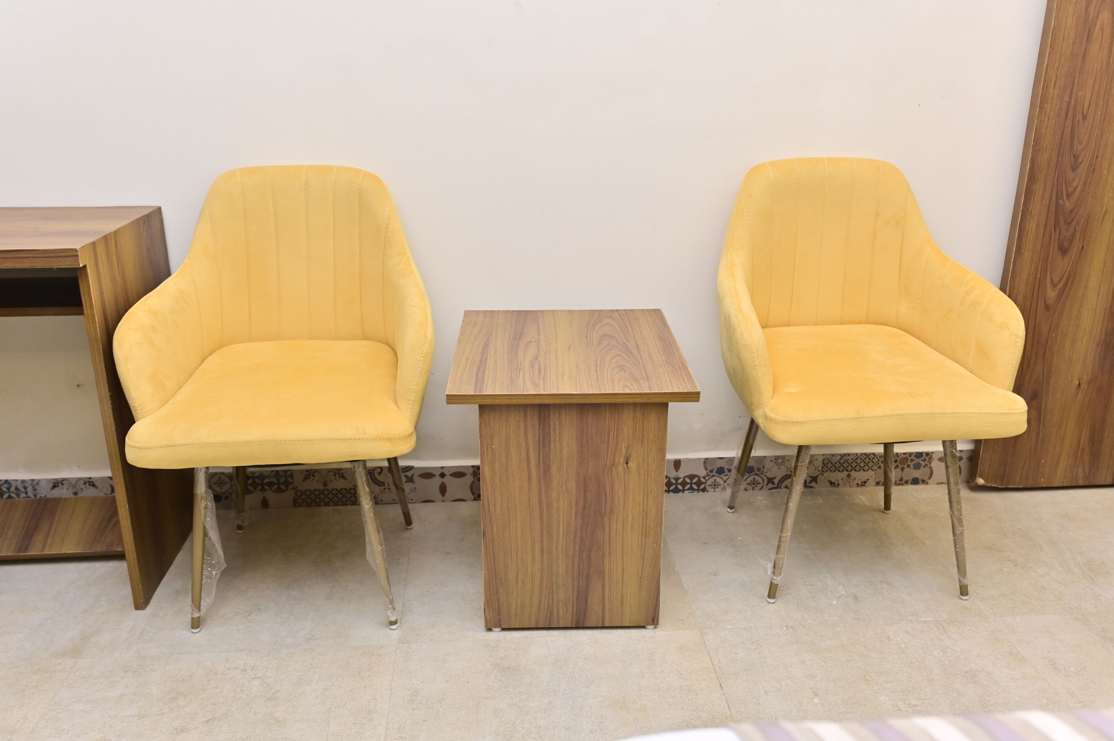 Two yellow chairs and a small wooden table in a room with tiled flooring and a plain wall.