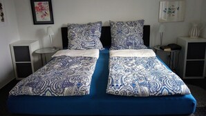 Un lit double avec une literie à motifs bleus et blancs, deux tables de chevet avec des lampes, une œuvre d’art florale encadrée accrochée au mur, et une petite table avec un vase et un livre.